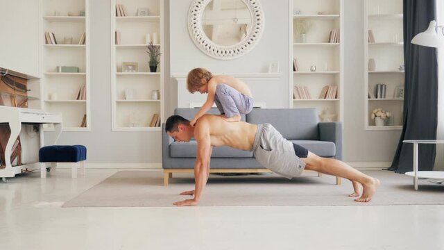 Sport time at home in the living room dad with his small cute son doing push-ups together while the small boy are on his back