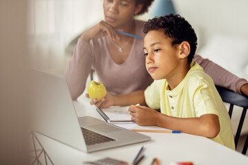 Little black boy using laptop while homeschooling with his mother.