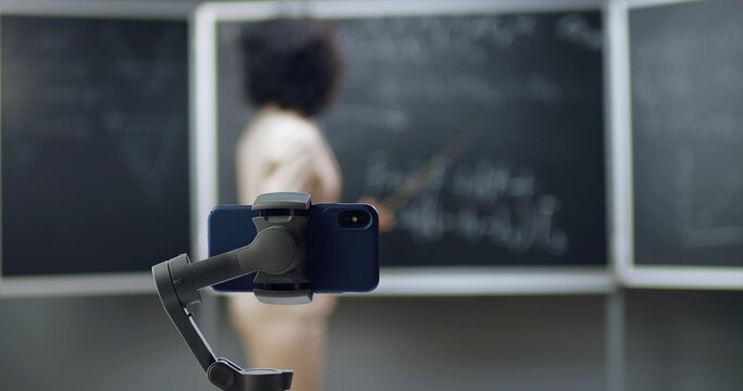 Smartphone recording video virtual lesson at school. Online studying. African American woman teacher explaining math or physics formulas at class on blackboard. Quarantine concept. Pandemic study.