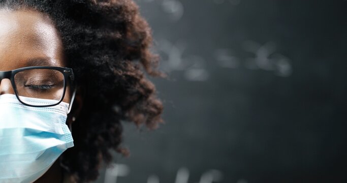 Close up of half face of young African American female teacher in glasses and medical mask looking at camera. Blackboard with math and physics formulas on background. School during pandemia. Portrait.