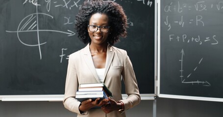 Portrait of young African American female teacher in glasses looking at camera in classrom and...