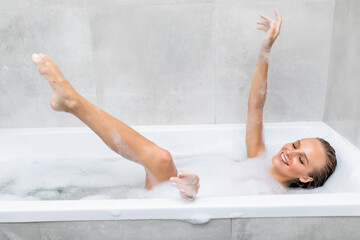 Happy laughing woman shaving her legs and having fun while lying in bathtub
