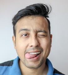 a young indian male winking with tongue out of mouth and a funny mischievous naughty expression on face isolated on grey with copy space for text.