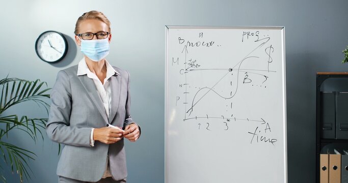 Caucasian young woman teacher in medical mask at school writing formulas and mahematics laws on blackboard. School concept. Female math lecturer in glasses explaining physics laws. Education. Covid-19