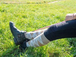 Crossed legs wear riding paddock boots and long knee socks. Horse rider hold wisp while sit in paddock of horse riding club.