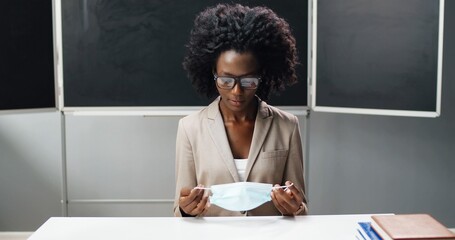 Portrait of African American female teacher in glasses taking on medical mask , sitting at table in classroom at school and smiling to camera. Woman lecturer wearing protection. Coronavirus concept.