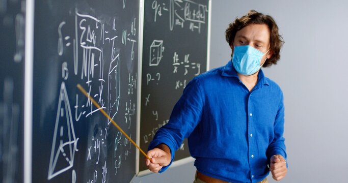 Caucasian male teacher in medical mask standing at board in classroom and telling physics or geometry laws to class. Pandemic concept. School during coronavirus. Educative mathematics lection.