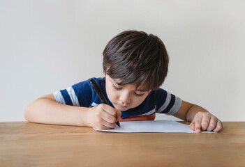 Naklejka premium Portrait of school kid sitting alone doing homework, Child boy holding black pen drawing and writing on white paper on table, Elementary school and homeschooling concept