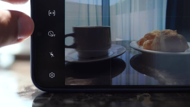 Closeup view 4k video of male hand holding modern smartphone. Man takes mobile photos and records video of tasty breakfast of his girlfriend. Cup of espresso coffee and two croissants on table.