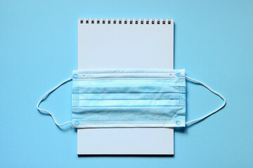 New normal concept with a blue face mask and white blank notebook page on light blue background