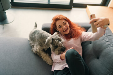Girl with red hair and charming smile lying on sofa and making selfie with her pet