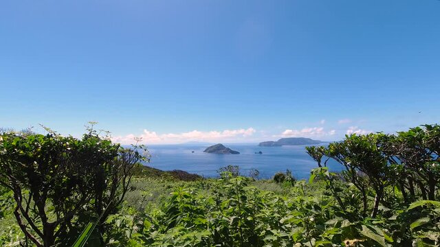 Wide view through greenery towards open ocean with islands and moving clouds on bright and sunny day - Time Lapse