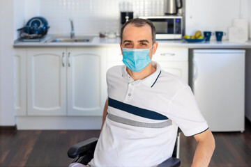 Smiling young handicapped man with medical mask sitting on wheelchair in kitchen in quarantine