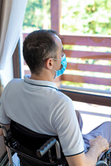 Young man wearing face mask sitting in a wheelchair alone looking out the window