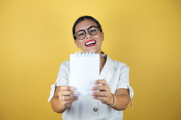 Beautiful woman over yellow background smiling and showing blank notebook in her hand