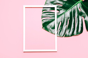 Empty picture frame with monstera leaves on pink background, top view