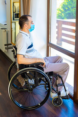 Young man wearing face mask sitting in a wheelchair alone looking out the window