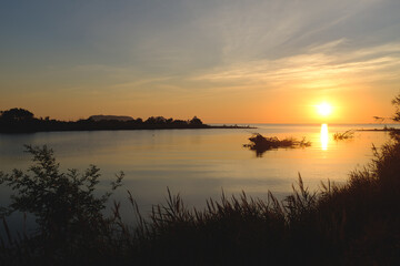 Fototapeta premium Orange sun rising on a shore coastal landscape with wild plants silhouette and sunlight reflecting on water
