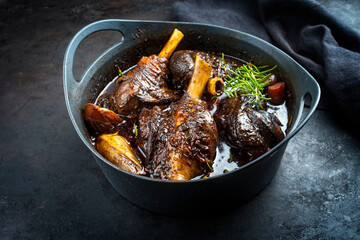 Modern style traditional braised slow cooked lamb shank in red wine sauce with shallots and carrots offered as close-up in a design stewpot