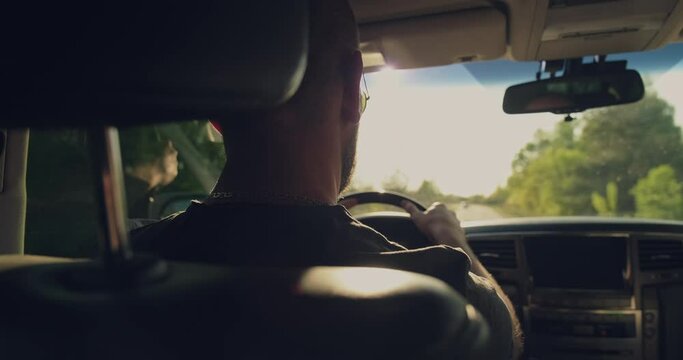 Rear View to the man drives a car. Man drives the car while sunset.  Young man driving the car.  Adult boy driving car. Real time. Sun's rays shine through the windshield of the car.
