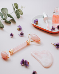 Obraz na płótnie Canvas Pink hyaluronic acid serum, rose quartz beauty roller, gua sha and flowers on pink table top. Anti aging cosmetics and tools, copy space, top view