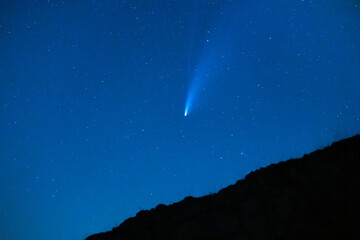 Trace of a comet or shooting star behind a hill in a blue starry sky with stars at night.