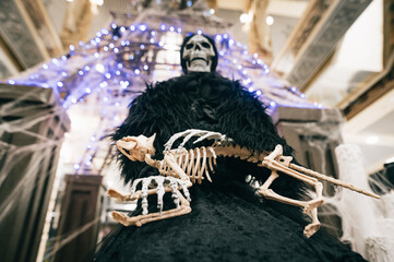 Death skeleton in black clothes stands indoors on Halloween with animal bones in hands. Halloween decorations. Background