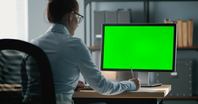 Concentrated woman in formal clothing sitting at private office and taking notes during video conference on computer with green screen. Concept of working process.
