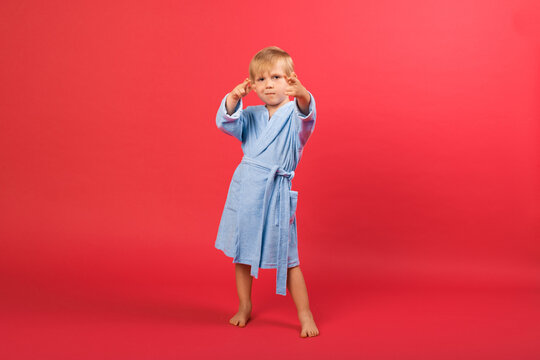 A Blond Child Boy In Blue Bathrobe On A Red Bright Background Shows Hand Sign Shoots Gun Or Aims Target Near Eyes. Studio Shot Of A Child Dancing In Bathhouse, Sauna, Spa, Pool And Swimming School.