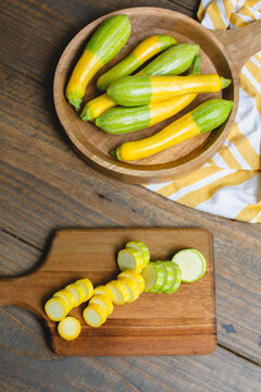 Top View of Fresh Zephyr Squash Sliced on a Wooden Cutting Board; Whole Squash in a Wooden Bowl on a Wooden Table in Background