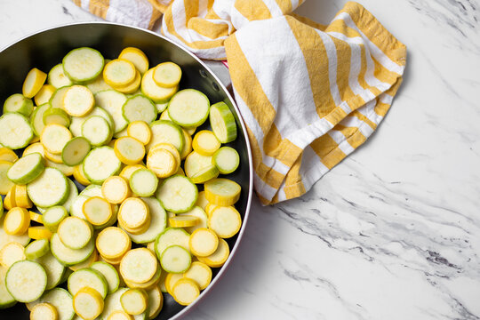 Top View of Sliced Zephyr Squash in Pan Ready to Cook; White Countertop with Yellow and White Kitchen Towel