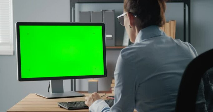 Back view of successful business woman sitting at office and typing on keyboard of computer with empty green screen. Space for text and advertisement.