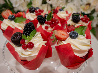 Delicious cupcakes with berries, fruit, whipped cream and mint leaves in red wrapping paper. Close-up, soft focus