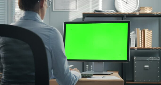Female company member in formal wear and eyeglasses having video call on computer with empty chroma key monitor. Business woman sitting at office during working online conference.