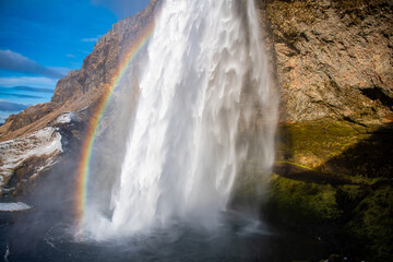 Waterfall from a cliff with a rainbow and mist in Iceland. The area behind the waterfall can be seen.