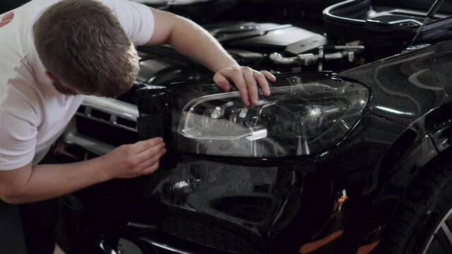 Crop mechanic cutting protective film on headlight. Ppaint protection film installation on front headlight of modern luxury car. Polyurethane film applied to car surface to protect the headlight from