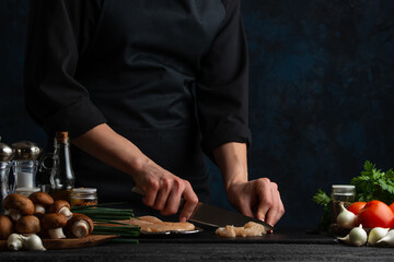 The chef in black uniform cutting the chicken by the knife on the black board with mushrooms and vegetables near isolated on dark background. Cooking the dinner. Food concept. Restaurant's recipe.
