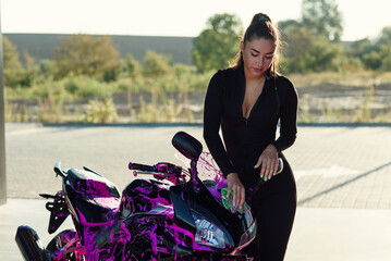 Fototapeta na wymiar Beautiful girl in tight-fitting seductive suit washes stylish sport motorcycle and wipes it from magenta foam at sunrise.