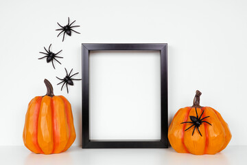Mock up black frame with pumpkin decor on a shelf or desk. Halloween concept. Portrait frame against a white wall with spiders.