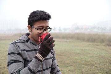 a man drinking hot coffee from a red cup and dressed in winter garments and gloves with focus on model's face with a foggy haze in the background