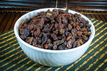 Raisins in a white bowl on a brown wooden mat.