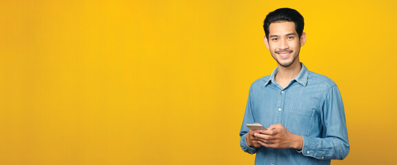 Asian man holding mobile phone smiling while standing isolated on yellow background with copy space...