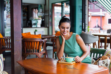Cute elegant calm happy woman in green summer dress sits with coffee in cafe enjoying morning. Beautiful coffee lover. 