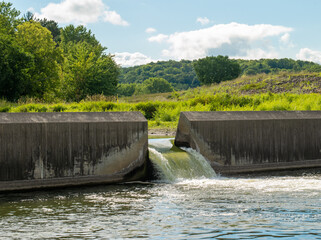 Water flows through the floodgate in a concrete dam on a river, with green trees and vegetation on the hills in the background.