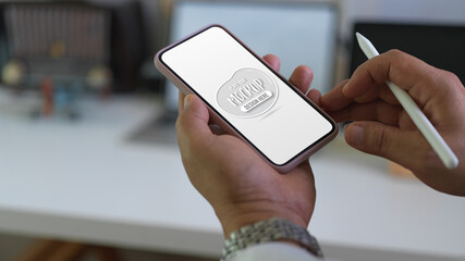 Male using mock up smartphone while holding stylus in blurred worktable background