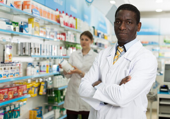Confident African American male pharmacist standing in interior of pharmacy .