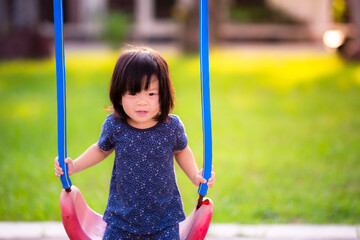 Concept of outside activities. Cute Asian little girls play swings on playground. Around playing field, there is a grass field as background. Cute and bright children. Sweet smile of young girl.