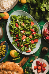 Turkish Mediterranean dinner table setting. Flat-lay of arugula, soft cheese and strawberry salad, rice pilav, flatbread and olives over oriental green tiled table background, top view. Turkish