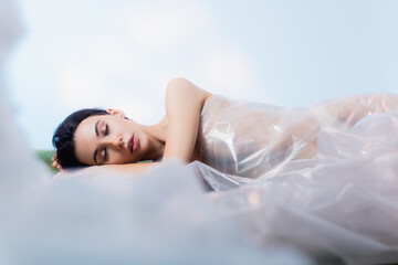 selective focus of brunette woman with closed eyes wrapped in polyethylene lying on blue, ecology...