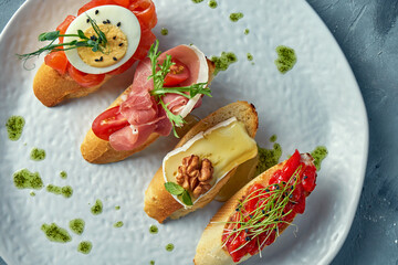 Assorted bruschetta with baked peppers, camembert, prosciutto and lightly salted salmon in a white plate on a gray background
